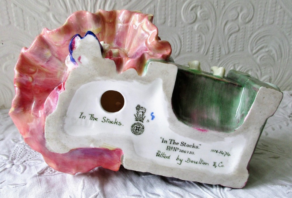 A hand-painted porcelain figurine depicting a woman in a ruffled pink dress seated alongside a small winged cupid trapped in green wooden stocks. View V