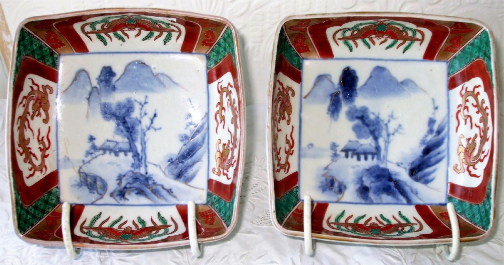 A pair of square Japanese Imari porcelain dishes featuring underglaze blue landscape scenes encircled by iron-red, green, and gold enamel borders.