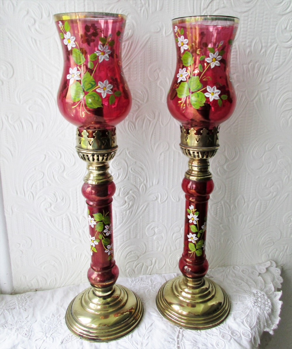 A pair of Victorian brass candle lamps featuring hand-painted floral enamel on red cranberry glass shades and stems.