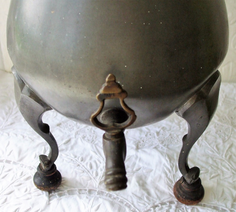 An antique pewter coffee urn featuring a pear-shaped body, an integrated tap at the front, a domed lid with a finial, and three cabriole legs terminating in turned wooden feet. View V
