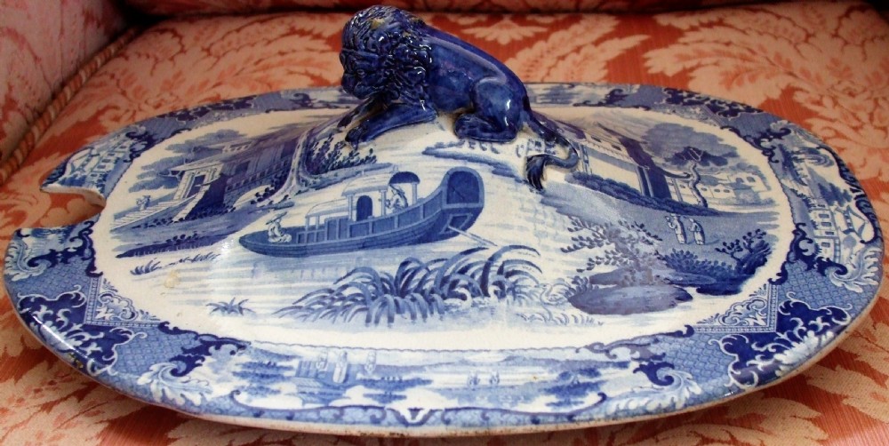 An oval blue and white transfer-printed ceramic tureen cover featuring a Chinoiserie river scene, a ladle notch, and a central lion finial handle. View II
