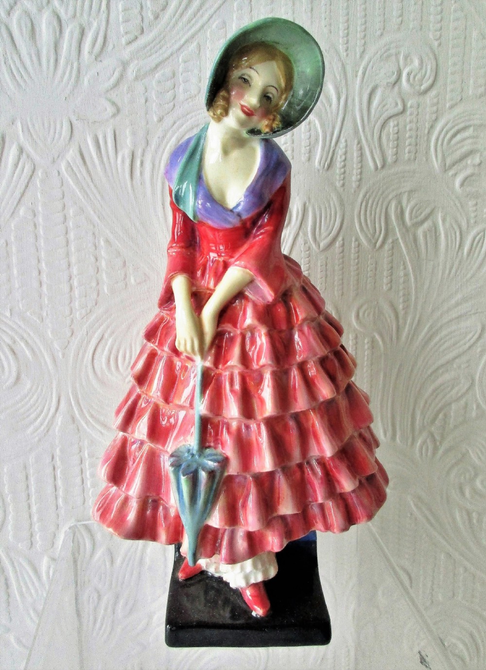 A Royal Doulton porcelain figurine titled Priscilla depicting a lady in a red tiered dress and green bonnet holding a parasol.