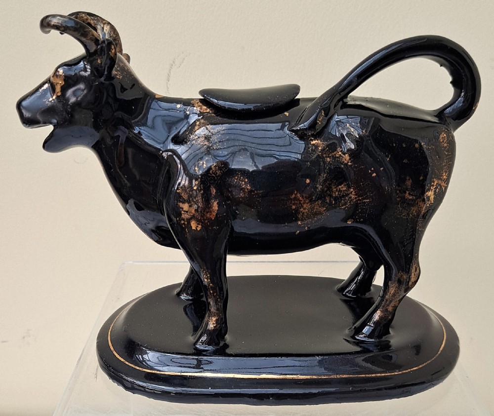 A Victorian Jackfield black-glazed ceramic cow creamer featuring gilded detailing and mounted on a contoured oval base.