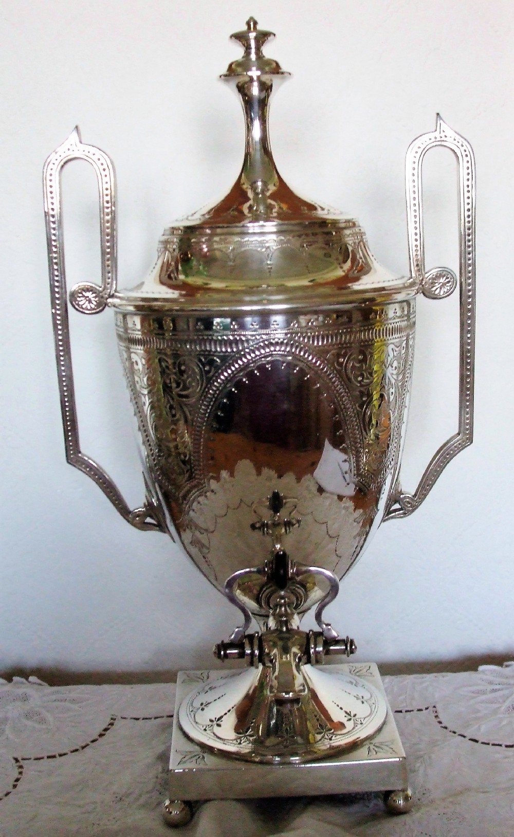 An Edwardian silver-plated samovar featuring engraved foliate motifs, high angular handles, a spigot tap, and a pedestal base resting on ball feet.