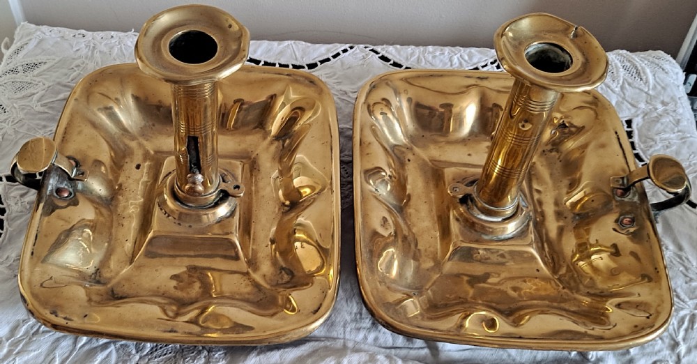 A pair of Victorian polished brass chambersticks with square dished trays, central socket columns, and side thumb handles.