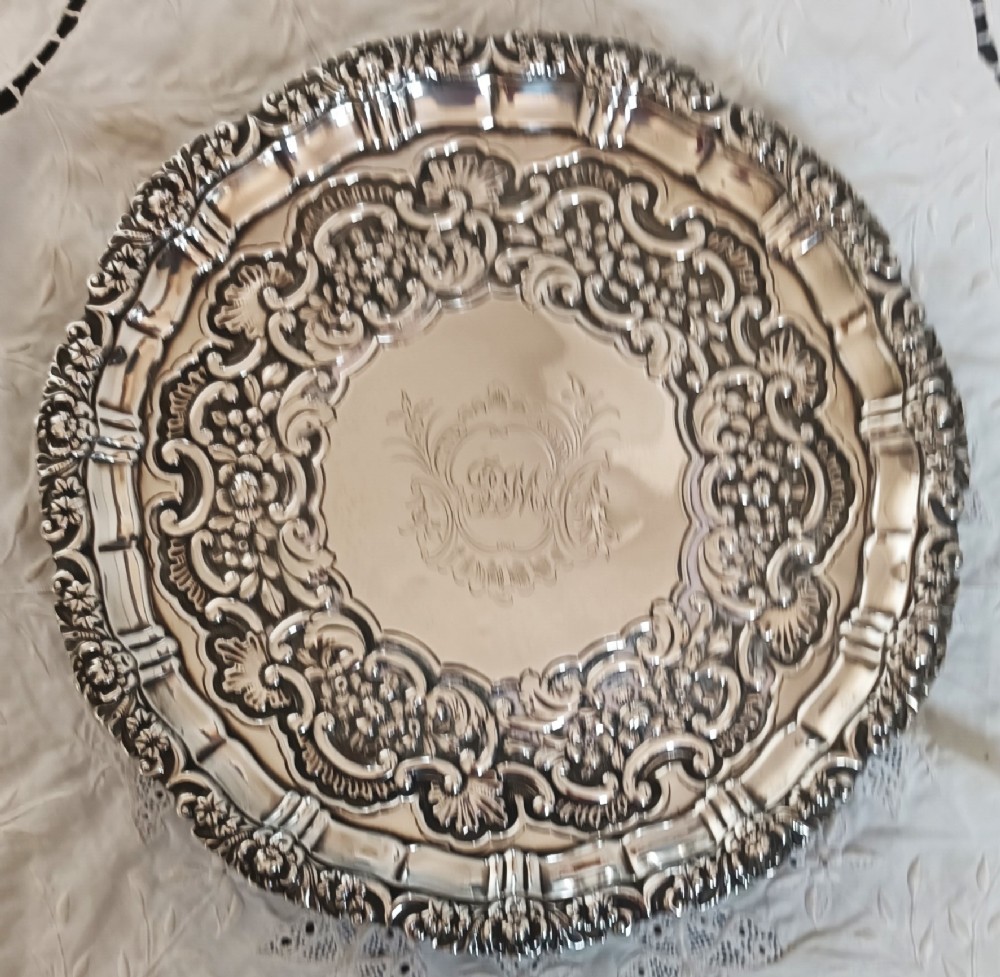 A circular Georgian sterling silver salver featuring an intricately chased scroll rim and a central engraved crest.