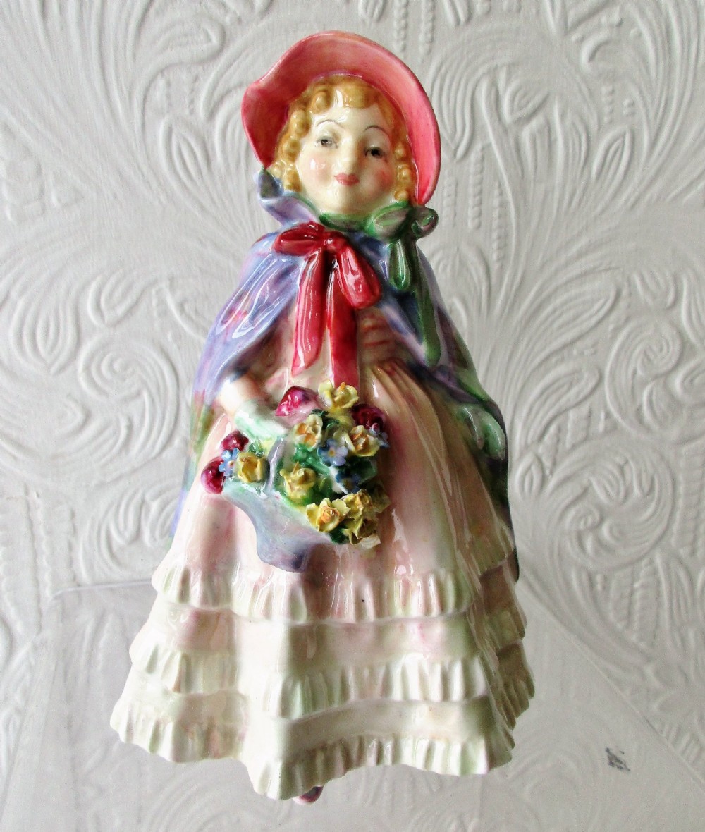 A hand-painted porcelain figurine of a girl wearing a pink bonnet, purple shawl, and layered ruffled dress while carrying a basket of flowers.