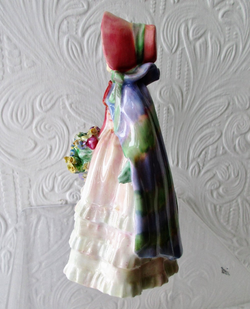 A hand-painted porcelain figurine of a girl wearing a pink bonnet, purple shawl, and layered ruffled dress while carrying a basket of flowers. View II