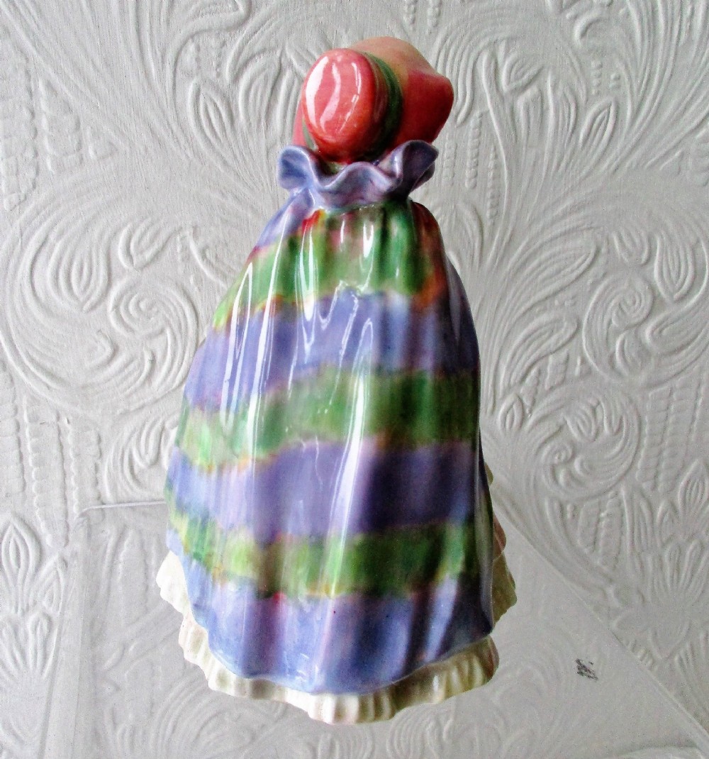 A hand-painted porcelain figurine of a girl wearing a pink bonnet, purple shawl, and layered ruffled dress while carrying a basket of flowers. View III