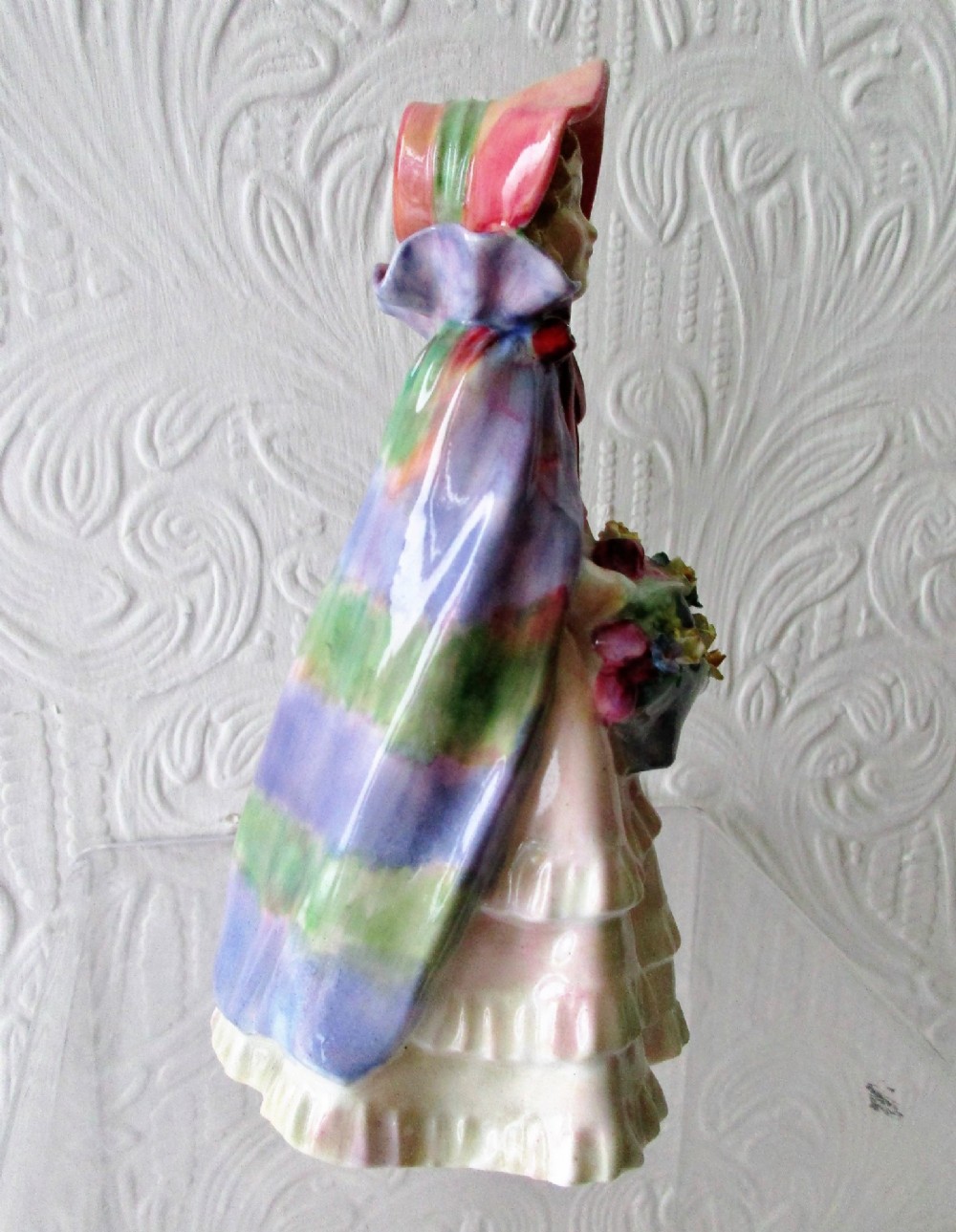 A hand-painted porcelain figurine of a girl wearing a pink bonnet, purple shawl, and layered ruffled dress while carrying a basket of flowers. View IV