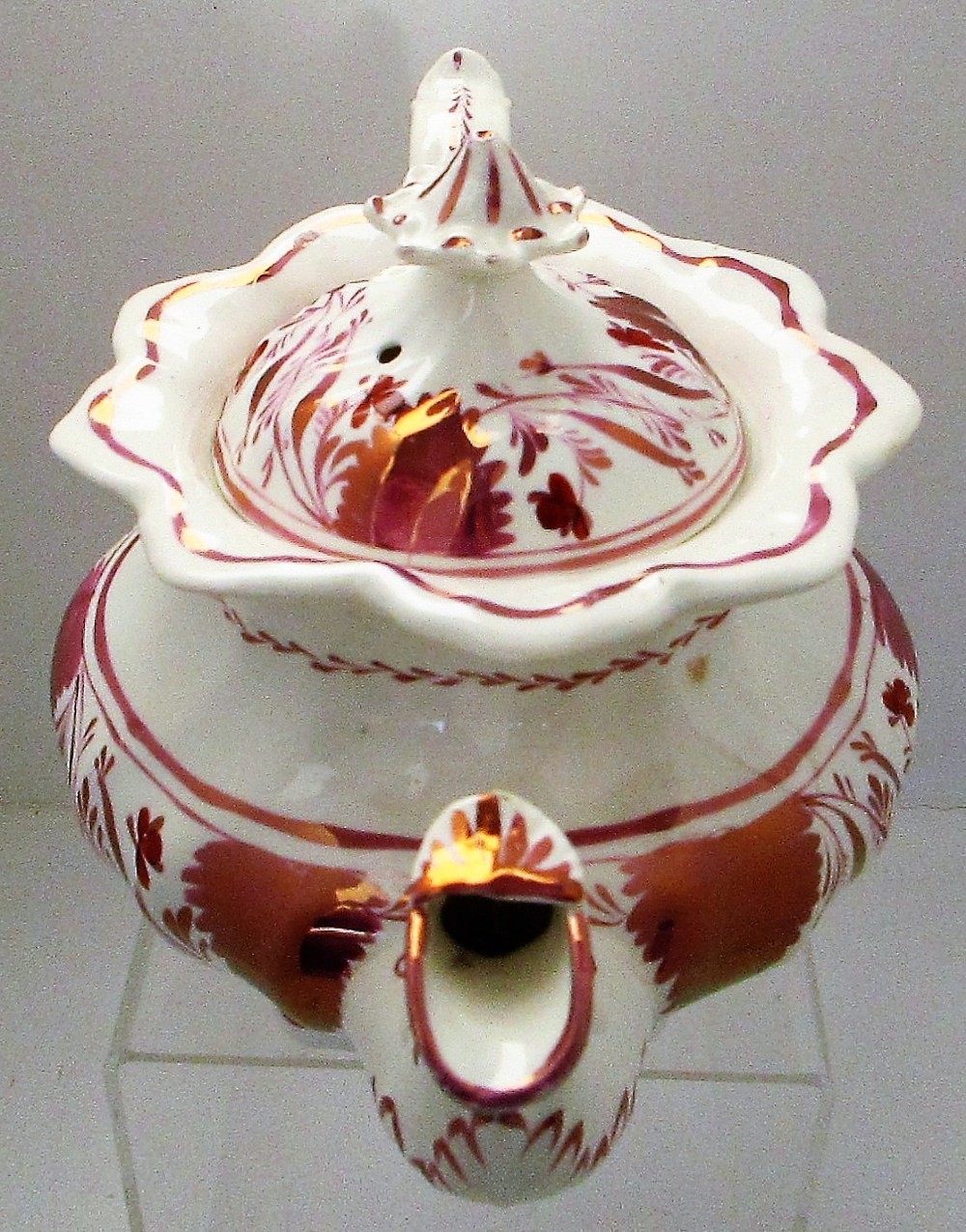 An antique white porcelain teapot decorated with pink iridescent metallic leaf motifs, a scalloped base, curved spout, and molded finial lid. View V