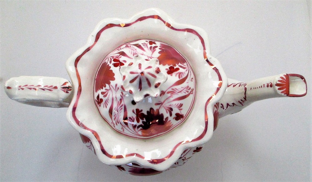 An antique white porcelain teapot decorated with pink iridescent metallic leaf motifs, a scalloped base, curved spout, and molded finial lid. View VI