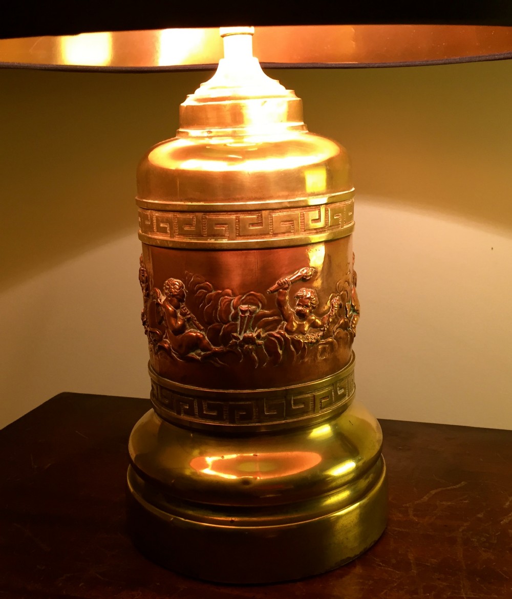 A cylindrical brass and copper table lamp featuring a central relief band depicting cherubs playing musical instruments alongside Greek key borders.