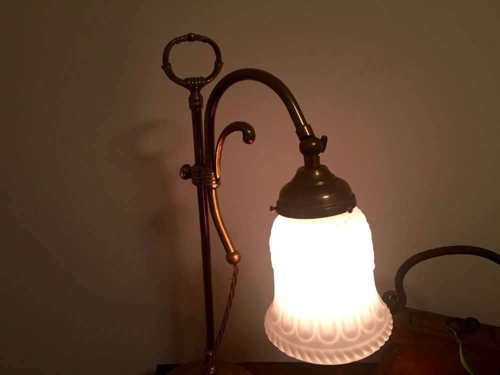 An Edwardian adjustable brass desk lamp featuring a curved arm, circular stepped base, top loop handle, and a illuminated white shade. View VI