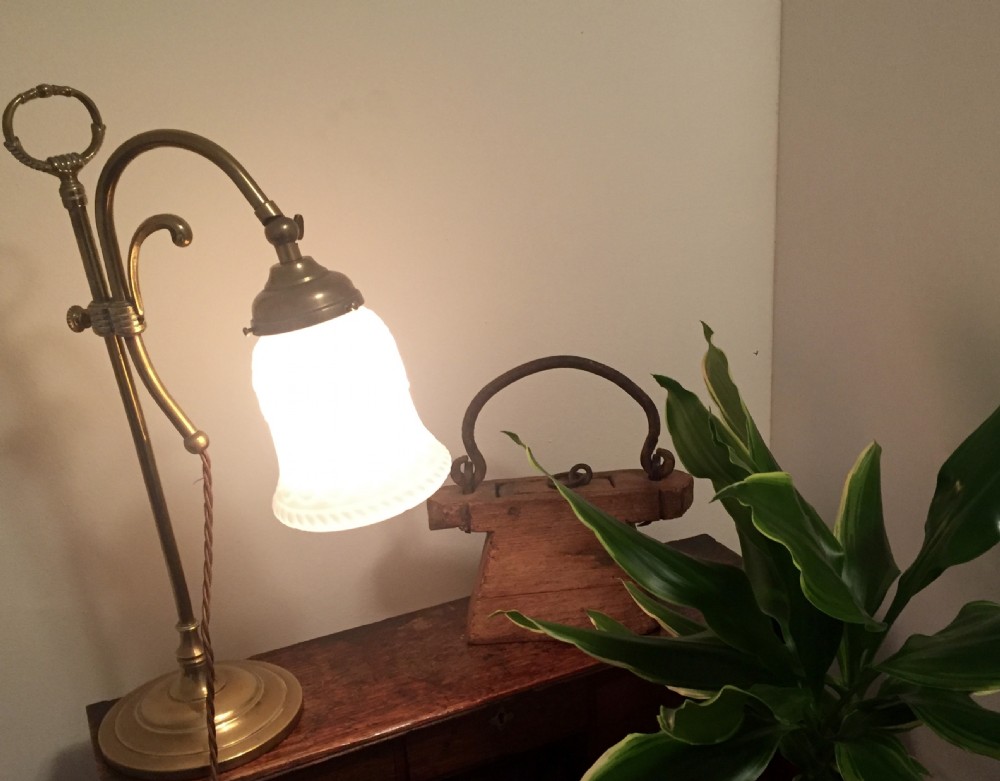 An Edwardian adjustable brass desk lamp featuring a curved arm, circular stepped base, top loop handle, and a illuminated white shade. View X