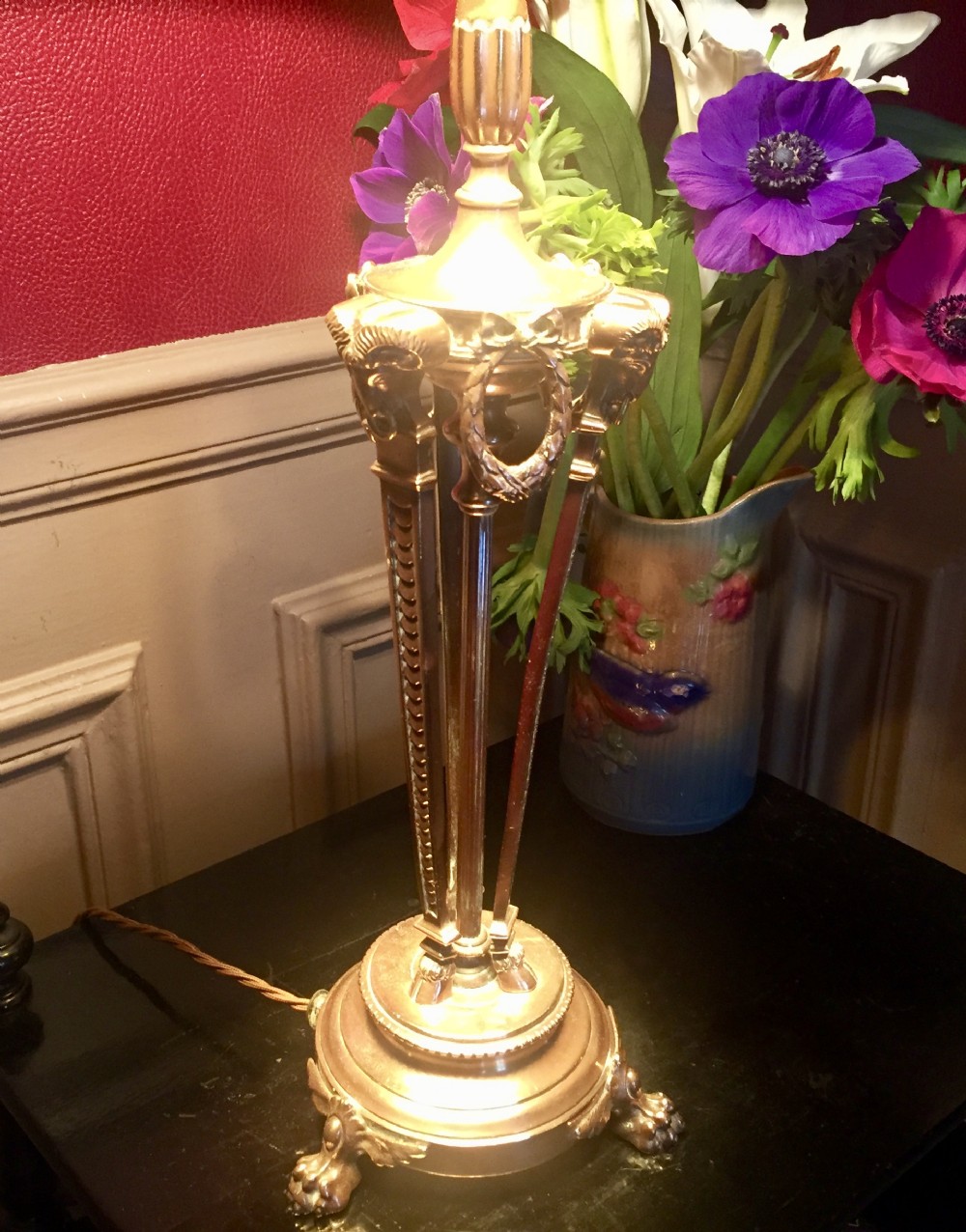 A French Empire style gilt metal table lamp base featuring three tripod legs decorated with rams heads, laurel wreaths, and claw feet.