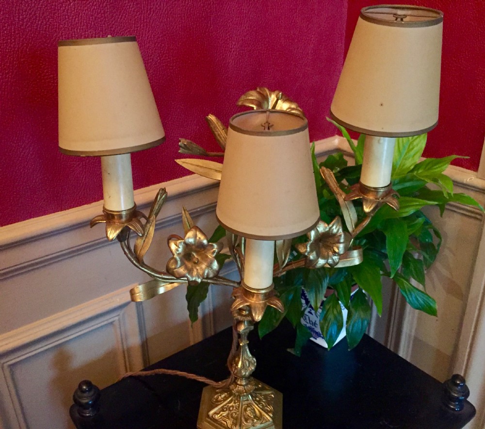 A 1920s French gilt brass three-arm candelabra table lamp featuring botanical leaf and blossom motifs with neutral fabric shades.