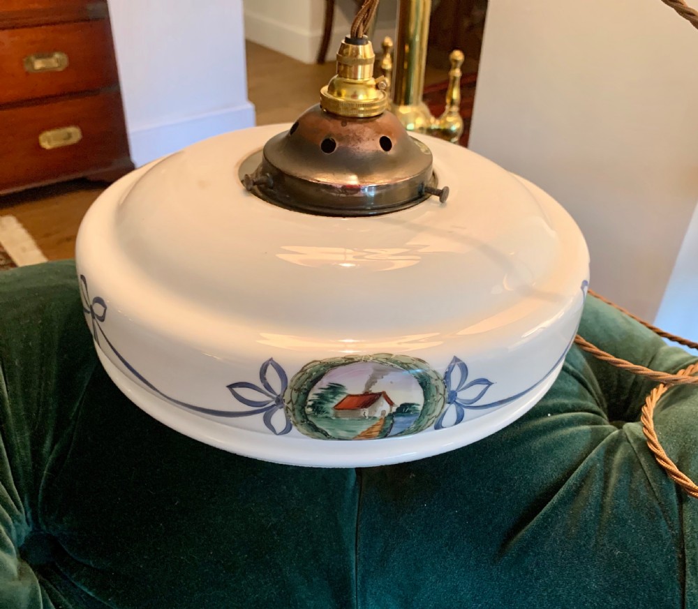 A white glass ceiling pendant shade with a brass top gallery, decorated with hand-painted blue ribbons and a central medallion depicting a cottage.