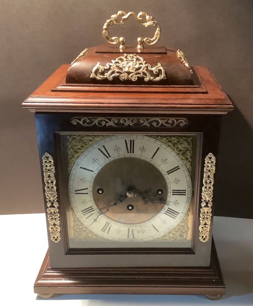 A dark mahogany bracket clock with a silvered Roman numeral dial, three winding holes, brass spandrels, ornate brass corner mounts, and a brass carrying handle.