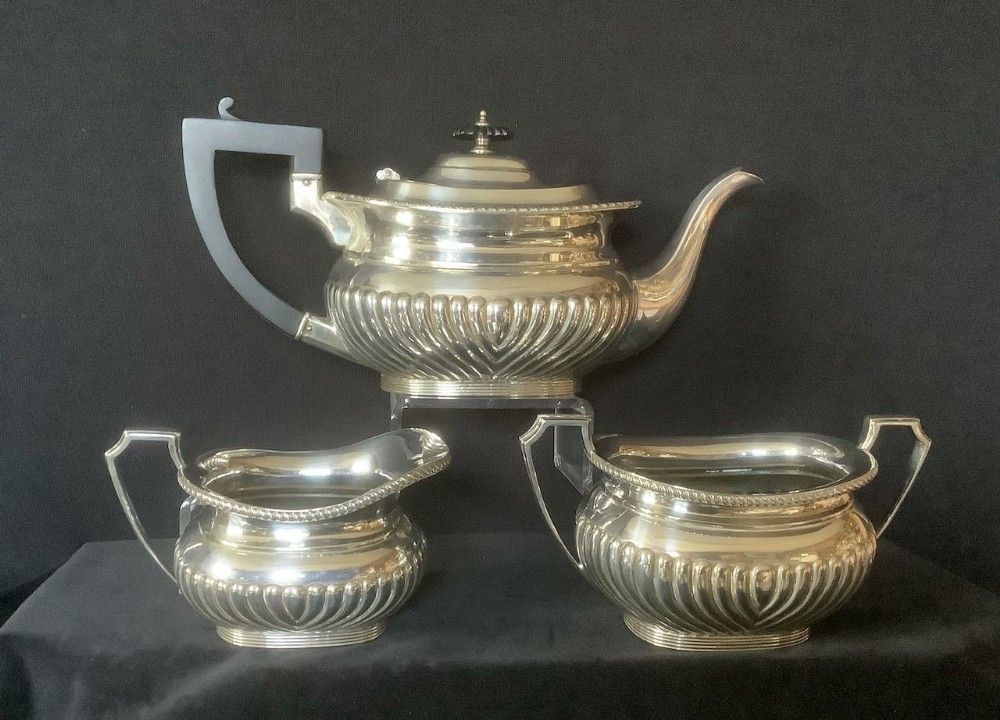 A three-piece silver-plated tea set featuring a teapot with an ebonised handle, a cream jug, and a sugar bowl, all with fluted gadrooned decoration.