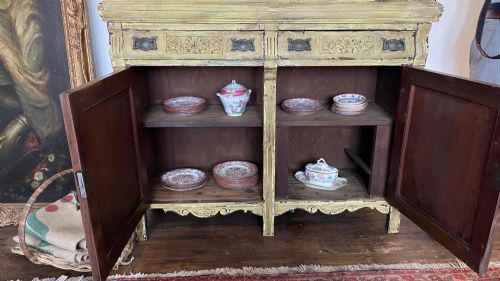 fig 4. An antique French provincial sideboard featuring distressed yellow painted wood, two carved panel doors with urn motifs, two upper drawers, and a pink marble top surface.