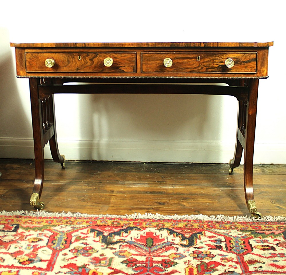 An antique Regency rosewood writing table featuring a rectangular top with rounded corners, a hinged stationery compartment, beaded lower rim, turned spindle uprights, and curved legs on brass feet. View III