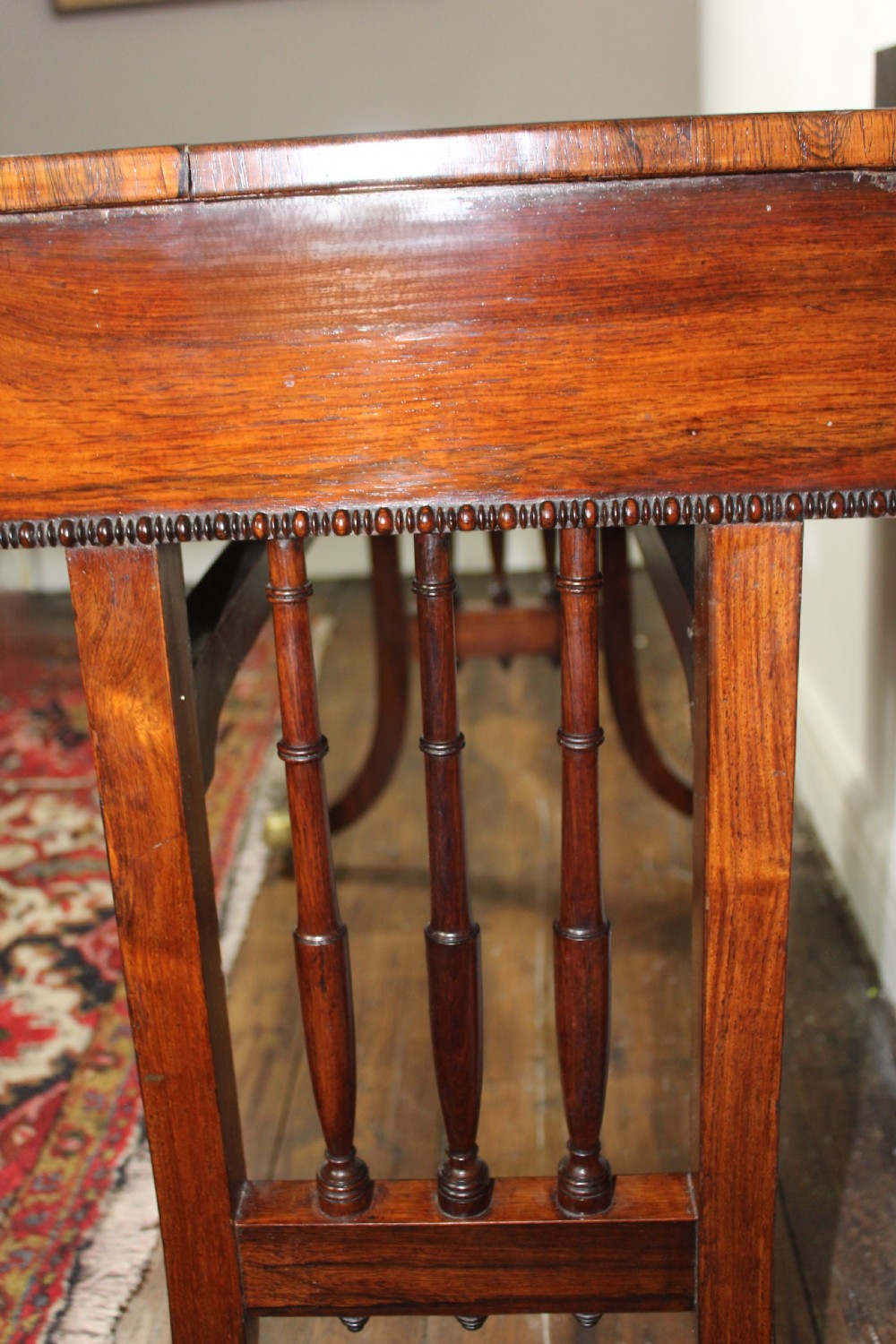 An antique Regency rosewood writing table featuring a rectangular top with rounded corners, a hinged stationery compartment, beaded lower rim, turned spindle uprights, and curved legs on brass feet. View V
