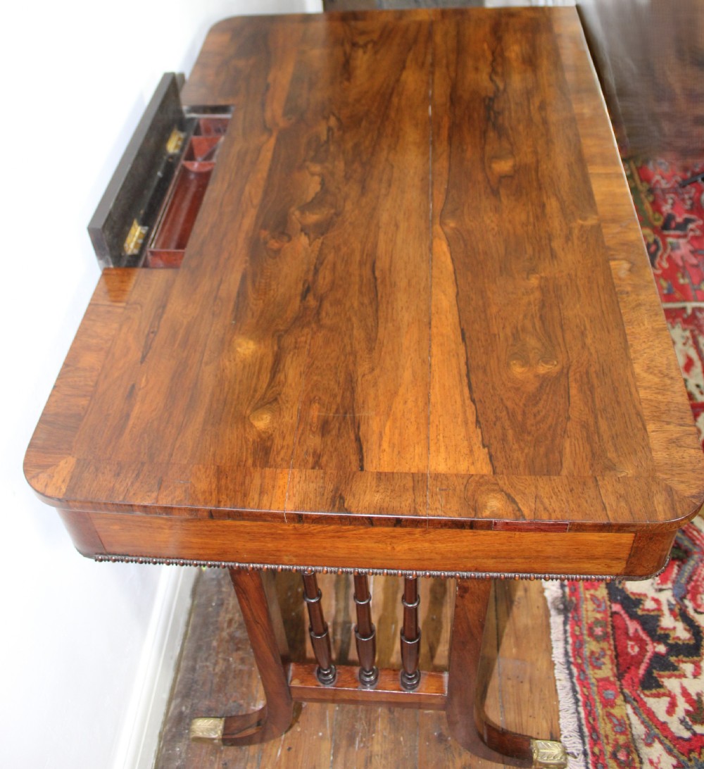An antique Regency rosewood writing table featuring a rectangular top with rounded corners, a hinged stationery compartment, beaded lower rim, turned spindle uprights, and curved legs on brass feet. View VI