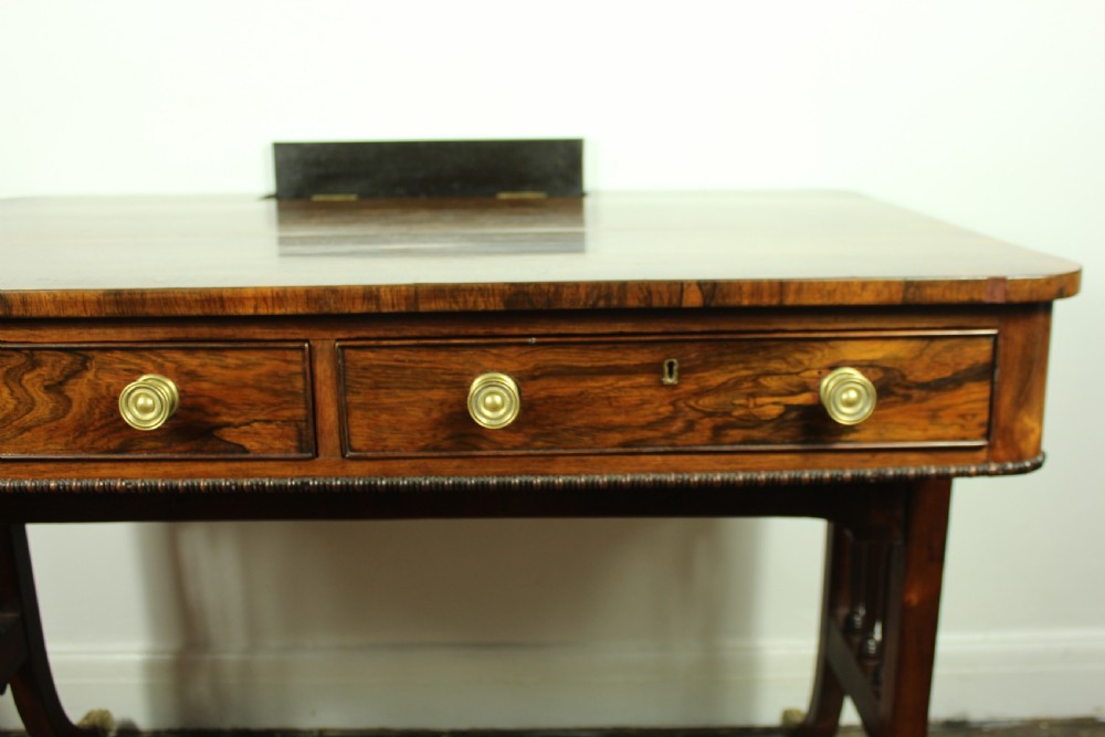 An antique Regency rosewood writing table featuring a rectangular top with rounded corners, a hinged stationery compartment, beaded lower rim, turned spindle uprights, and curved legs on brass feet. View VII