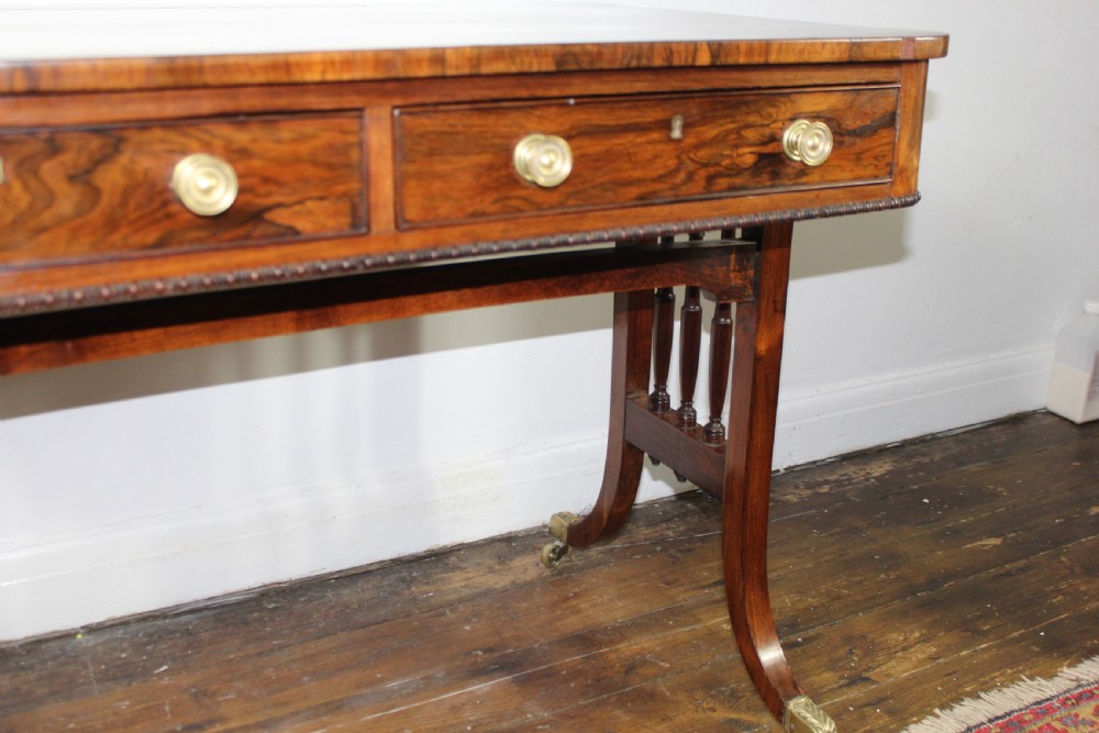 An antique Regency rosewood writing table featuring a rectangular top with rounded corners, a hinged stationery compartment, beaded lower rim, turned spindle uprights, and curved legs on brass feet. View VIII