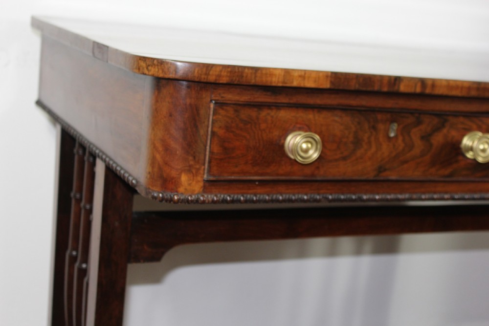 An antique Regency rosewood writing table featuring a rectangular top with rounded corners, a hinged stationery compartment, beaded lower rim, turned spindle uprights, and curved legs on brass feet. View IX