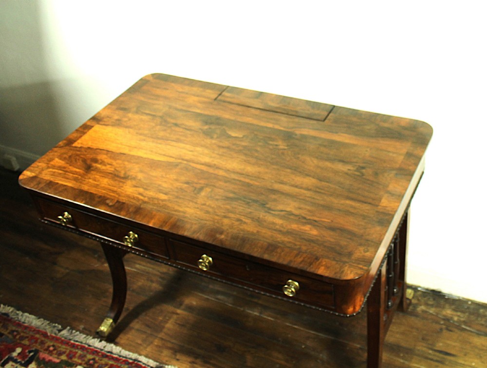 An antique Regency rosewood writing table featuring a rectangular top with rounded corners, a hinged stationery compartment, beaded lower rim, turned spindle uprights, and curved legs on brass feet. View X