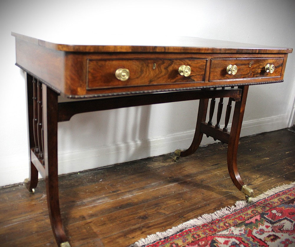 An antique Regency rosewood writing table featuring a rectangular top with rounded corners, a hinged stationery compartment, beaded lower rim, turned spindle uprights, and curved legs on brass feet. View XI