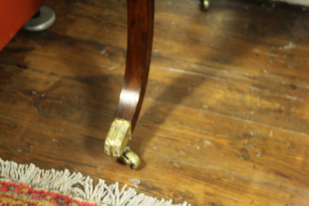 An antique Regency rosewood writing table featuring a rectangular top with rounded corners, a hinged stationery compartment, beaded lower rim, turned spindle uprights, and curved legs on brass feet. View XII