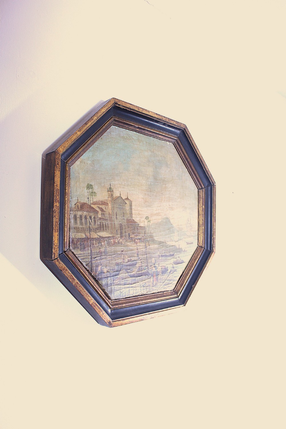 An octagonal framed early 19th-century oil painting depicting a Brazilian coastal landscape with colonial buildings, figures along the shore, and boats on the water. View VI