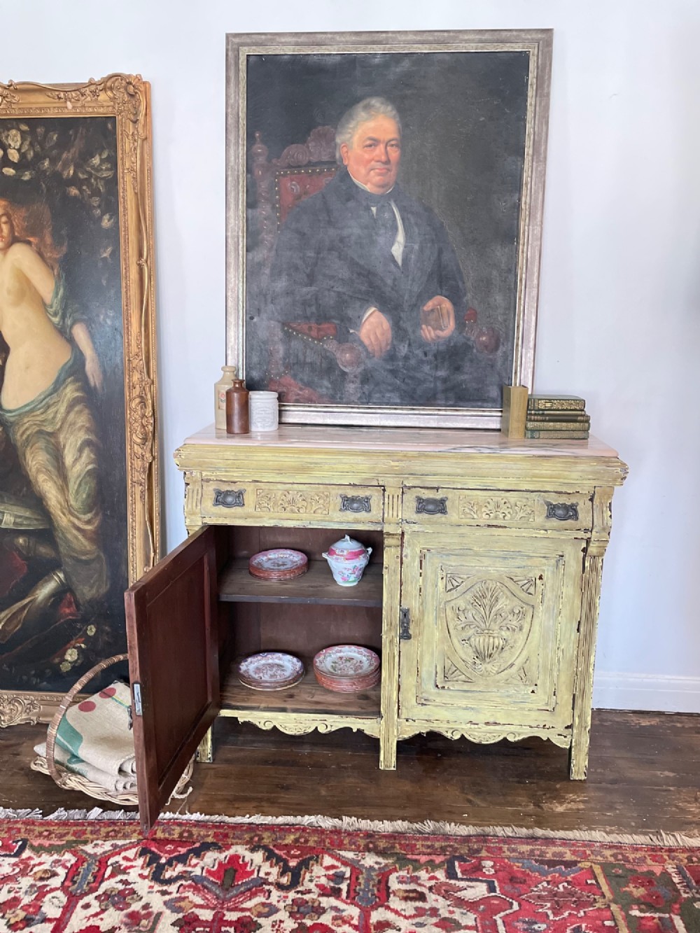 An antique French provincial sideboard featuring distressed yellow painted wood, two carved panel doors with urn motifs, two upper drawers, and a pink marble top surface. View II