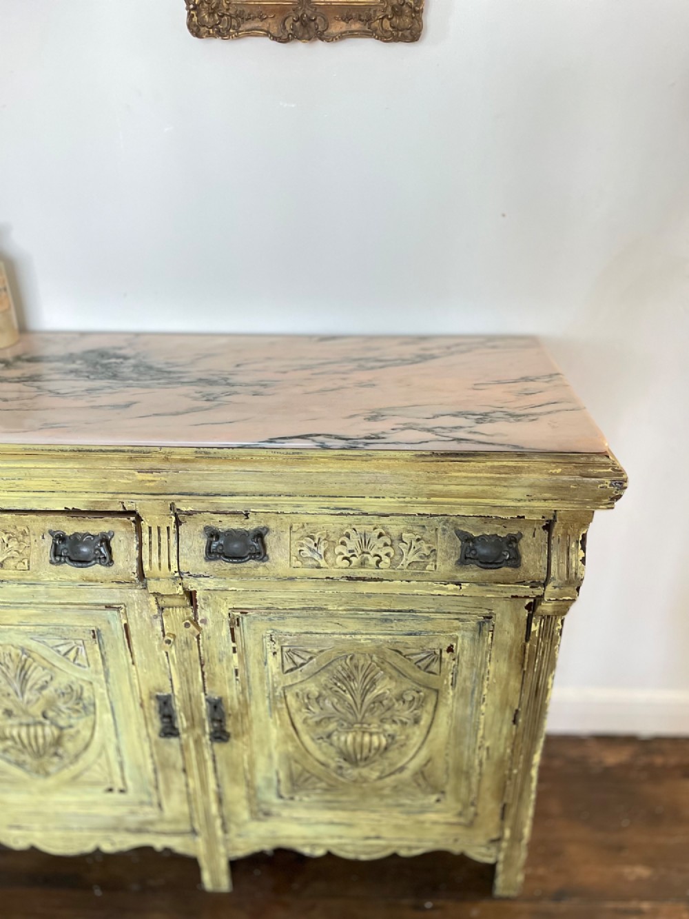 An antique French provincial sideboard featuring distressed yellow painted wood, two carved panel doors with urn motifs, two upper drawers, and a pink marble top surface. View XIII