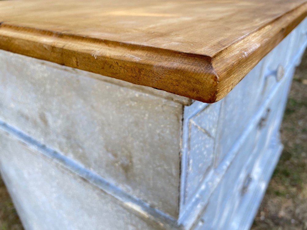 A French Gustavian style chest of drawers featuring a natural wood top, pale blue distressed paint finish, fluted corner pilasters, carved diamond-point details, and metal drop handles. View VI