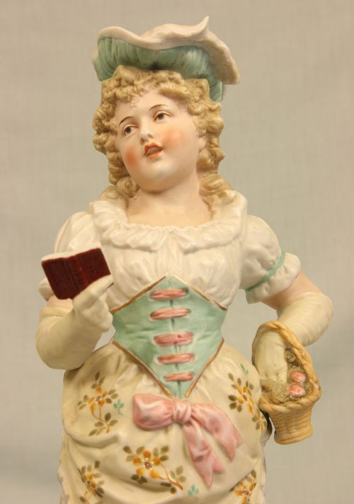 fig 1. An antique hand-painted bisque porcelain figurine of a young girl in Victorian dress holding a book and a basket.