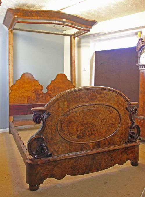 fig 1. An antique Victorian burr walnut half tester bed featuring a carved footboard with scrollwork and a fabric-lined overhead canopy.