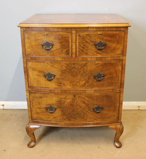 fig 1. A compact burr walnut chest of drawers featuring a bow front facade, brass drop handles, and cabriole legs with carved shell knees.
