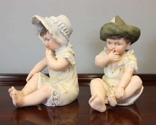 fig 1. A pair of antique bisque porcelain piano baby figurines depicting seated infants wearing bonnets and lace-trimmed garments with gilded accents.