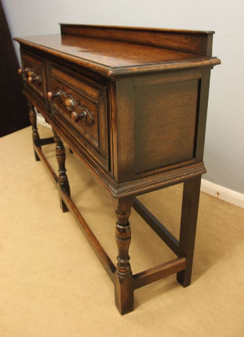 An antique dark oak side table with two geometric-panelled drawers, turned legs, a low stretcher base, and a raised backboard. View III
