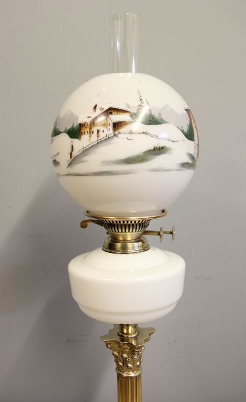 An antique oil lamp featuring a stepped brass Corinthian column base, a white glass font, a clear glass chimney, and a spherical white glass shade painted with a winter landscape. View IV