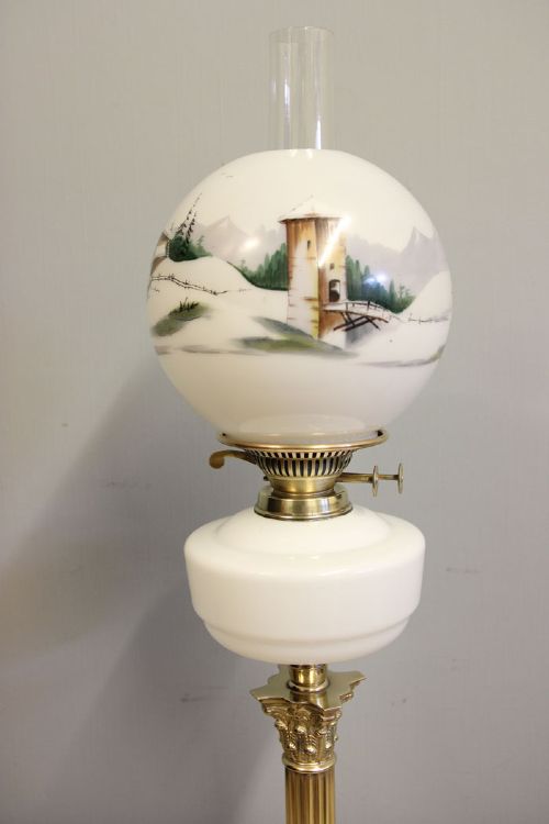 An antique oil lamp featuring a stepped brass Corinthian column base, a white glass font, a clear glass chimney, and a spherical white glass shade painted with a winter landscape. View VI