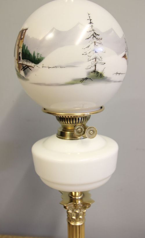 An antique oil lamp featuring a stepped brass Corinthian column base, a white glass font, a clear glass chimney, and a spherical white glass shade painted with a winter landscape. View VIII