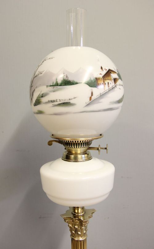 An antique oil lamp featuring a stepped brass Corinthian column base, a white glass font, a clear glass chimney, and a spherical white glass shade painted with a winter landscape. View XII