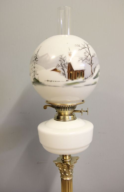 An antique oil lamp featuring a stepped brass Corinthian column base, a white glass font, a clear glass chimney, and a spherical white glass shade painted with a winter landscape. View XIII