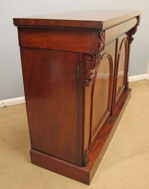 An antique Victorian flame mahogany chiffonier sideboard with two arched panel doors, carved corbels, and a plinth base. View V