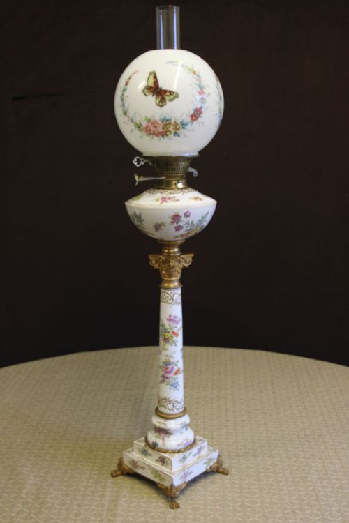 fig 1. An antique Victorian porcelain table oil lamp featuring hand-painted floral motifs, a butterfly printed globe shade, a brass Corinthian capital, and a square footed base.